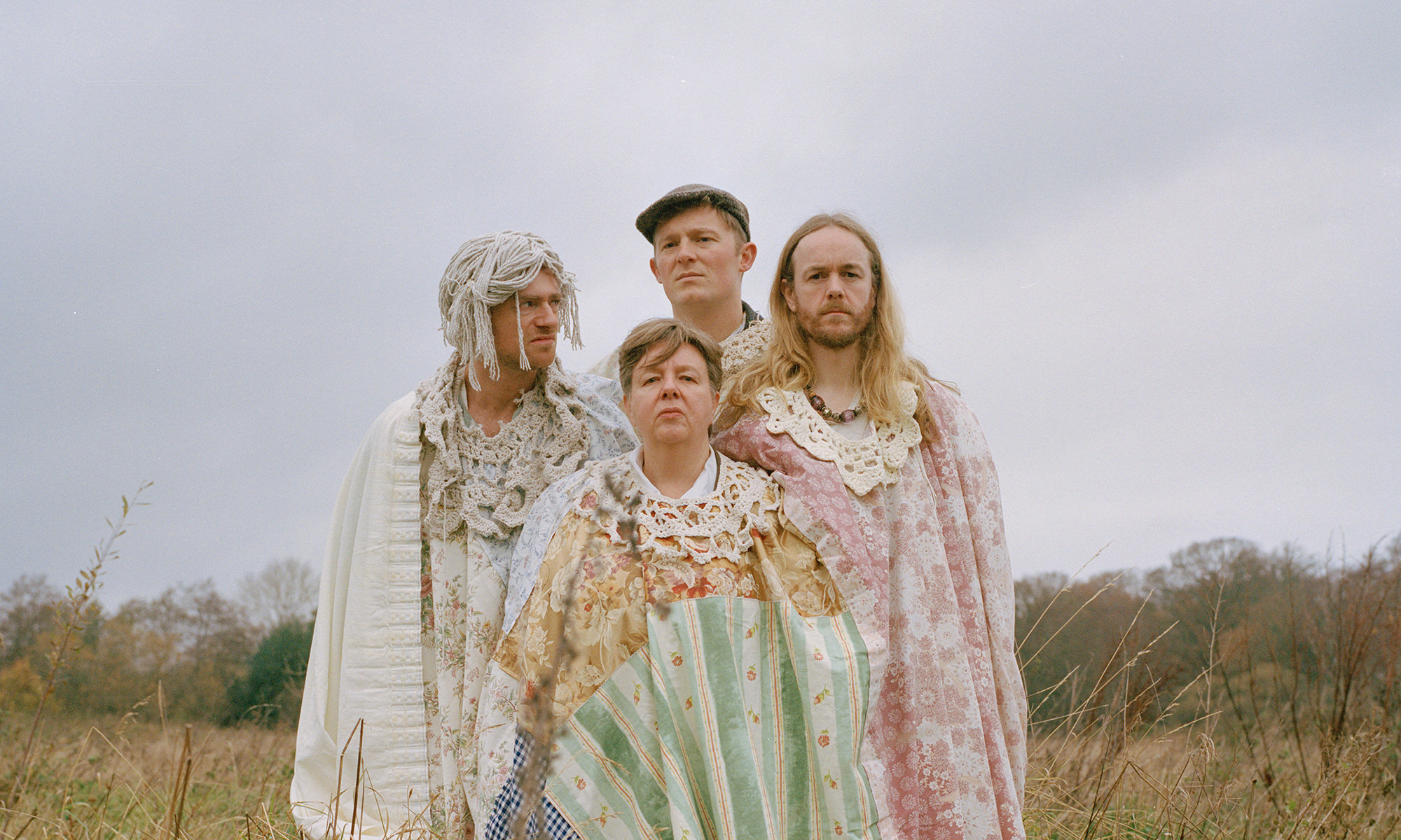 The four members of Bastard Assignments stand in the middle of a field, wearing a shared garment made out of a patchwork sheet, that drapes over their bodies. Their heads poke through holes in the sheet, their necks fringed with lacy collars. A grey cloudy sky and trees in the distance can be seen behind them